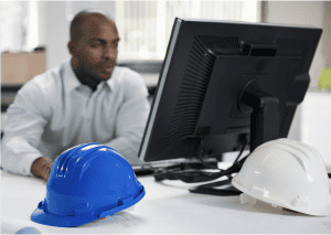man working on desktop with blue construction hat sitting on desk